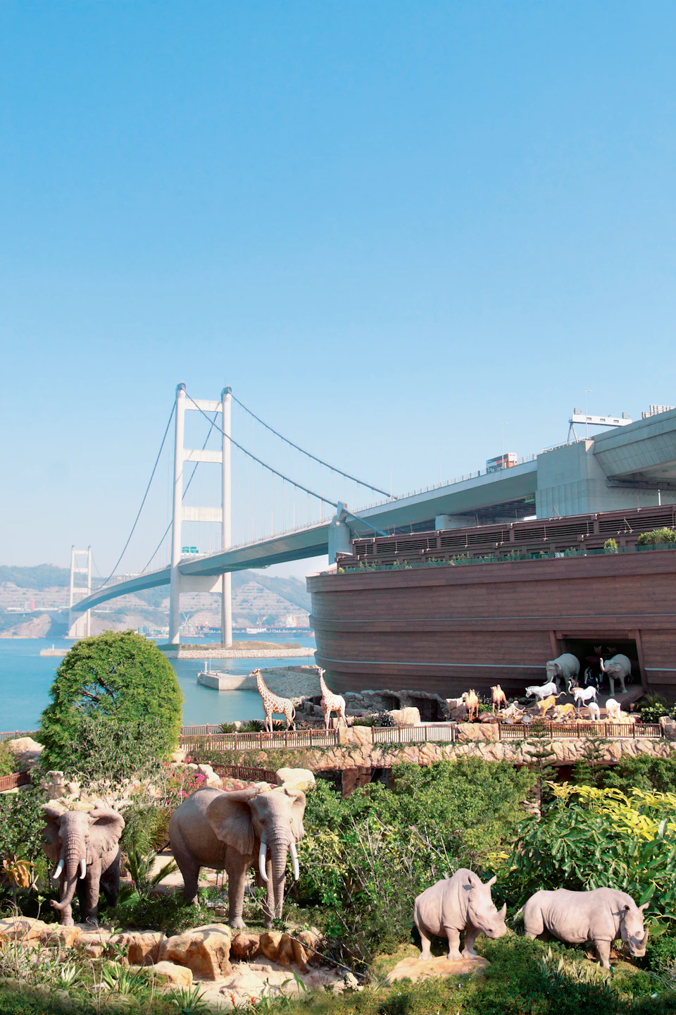 A large wooden building resembling Noah's Ark, surrounded by various animal sculptures, near a suspension bridge over water.