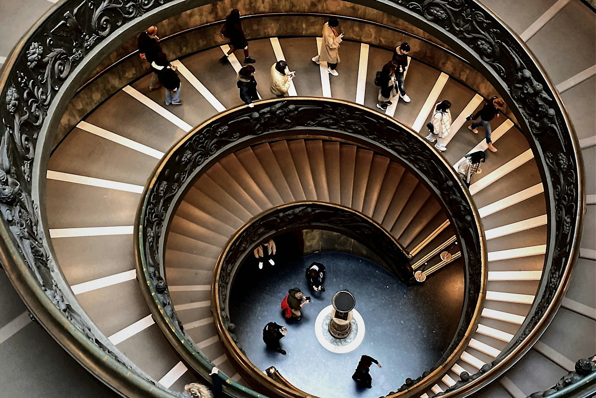 A spiral staircase with people walking on it, viewed from above, with ornate railings and a decorative column at the center.