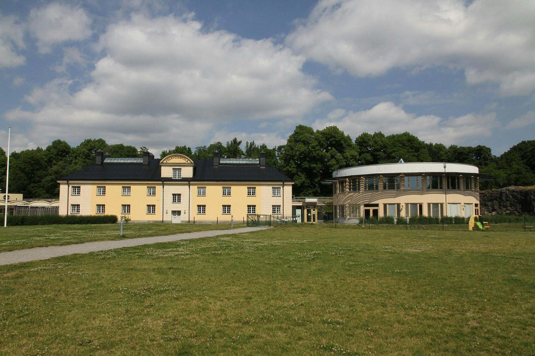 Path leading to a two-story yellow building with a black roof, adjacent to a round, modern structure. Grassy area in foreground.