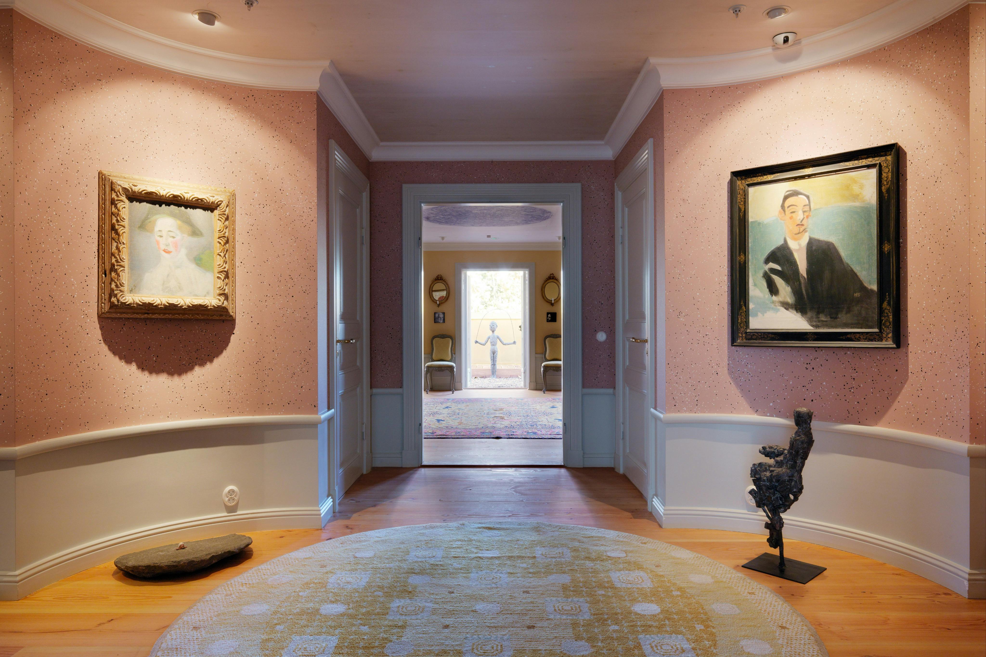 Corredor com chão de madeira, dois quadros emoldurados em paredes cor-de-rosa, um tapete redondo e uma escultura. A porta dá acesso a um quarto luminoso.