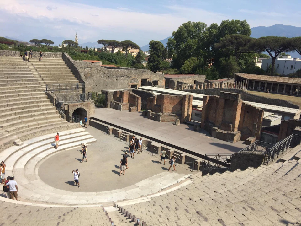 People explore the ancient ruins of an amphitheater with tiered seating, surrounded by trees and partially reconstructed walls.