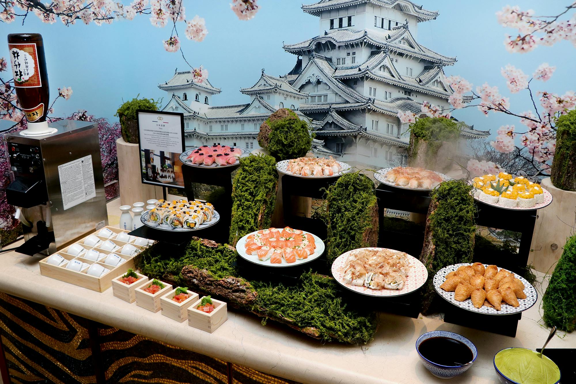 A decorative buffet setup featuring sushi and Japanese cuisine with a backdrop of a Japanese castle and cherry blossoms.