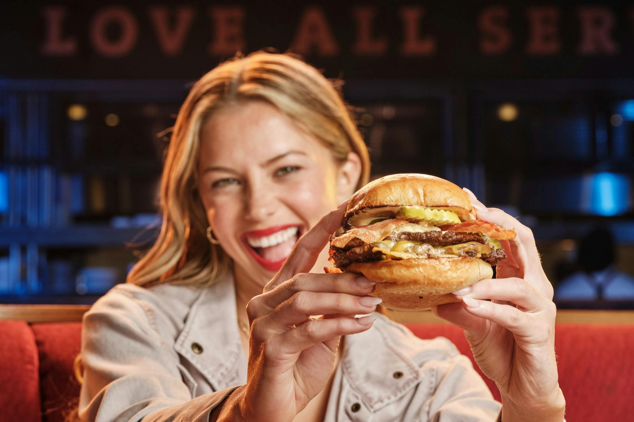 Žena, která se usmívá a drží hamburger v blízkosti kamery v restauraci s nápisem "LOVE ALL" v pozadí.