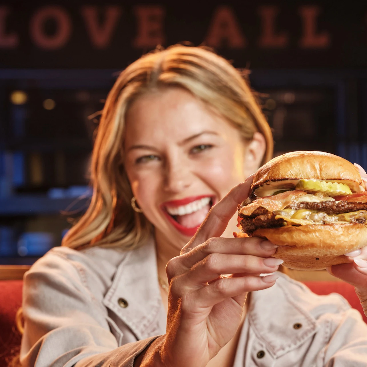 A woman smiling and holding a burger close to the camera in a restaurant with a sign that says 'LOVE ALL' in the background.