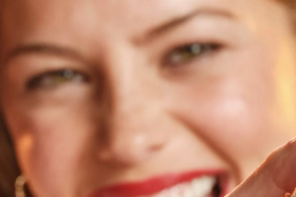 A woman smiling and holding a burger close to the camera in a restaurant with a sign that says 'LOVE ALL' in the background.