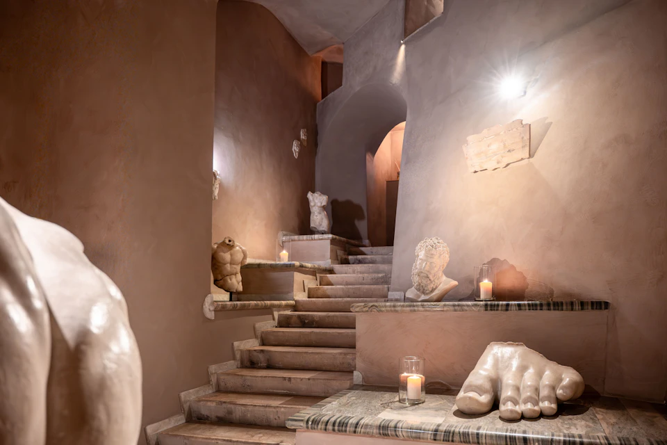 A dimly lit stone staircase with sculptures and candles placed on its steps and walls, creating an artistic ambiance.