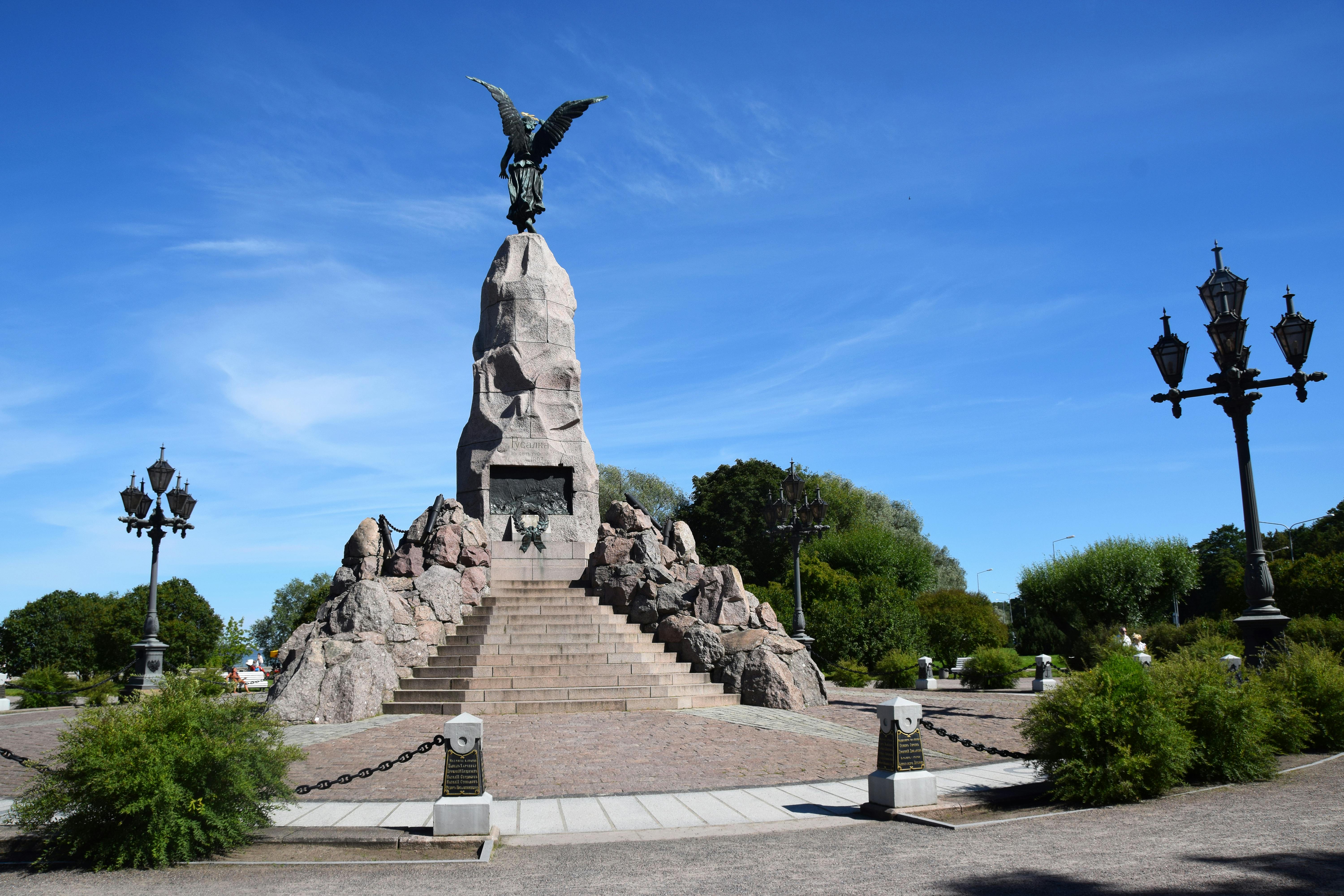 Stone monument with an eagle on top, surrounded by stairs and rocky structures, set in a landscaped park under a clear blue sky.