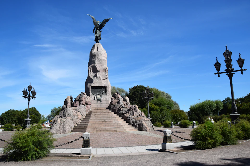 Stone monument with an eagle on top, surrounded by stairs and rocky structures, set in a landscaped park under a clear blue sky.