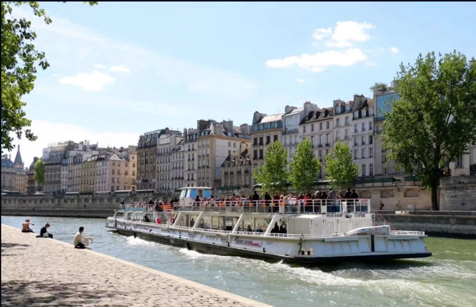 A tour boat with passengers cruises a river, bordered by historic buildings and trees under a partly cloudy sky.