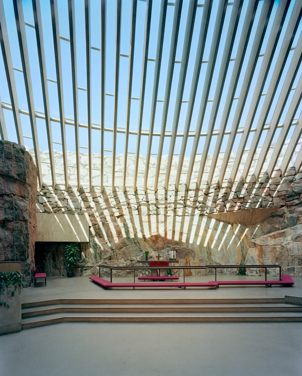 A spacious interior with rock walls and a skylight of angled beams, featuring benches and a table with candles on a tiered platform.