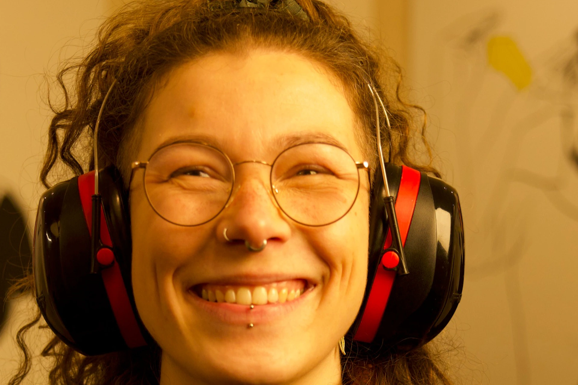 Two people wearing headphones smiling, with artwork displayed on the wall behind them in a warmly lit room.