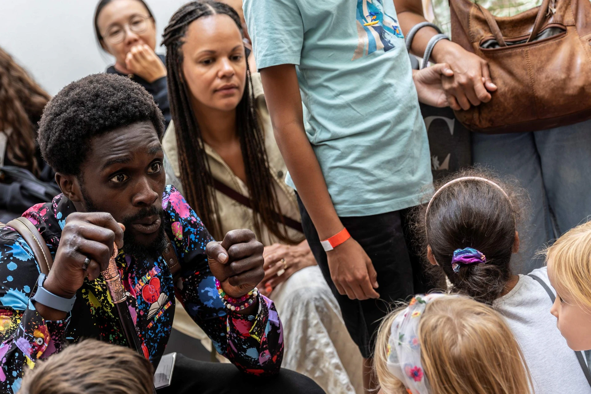 A man animatedly speaks to a group of children and adults gathered around him indoors.