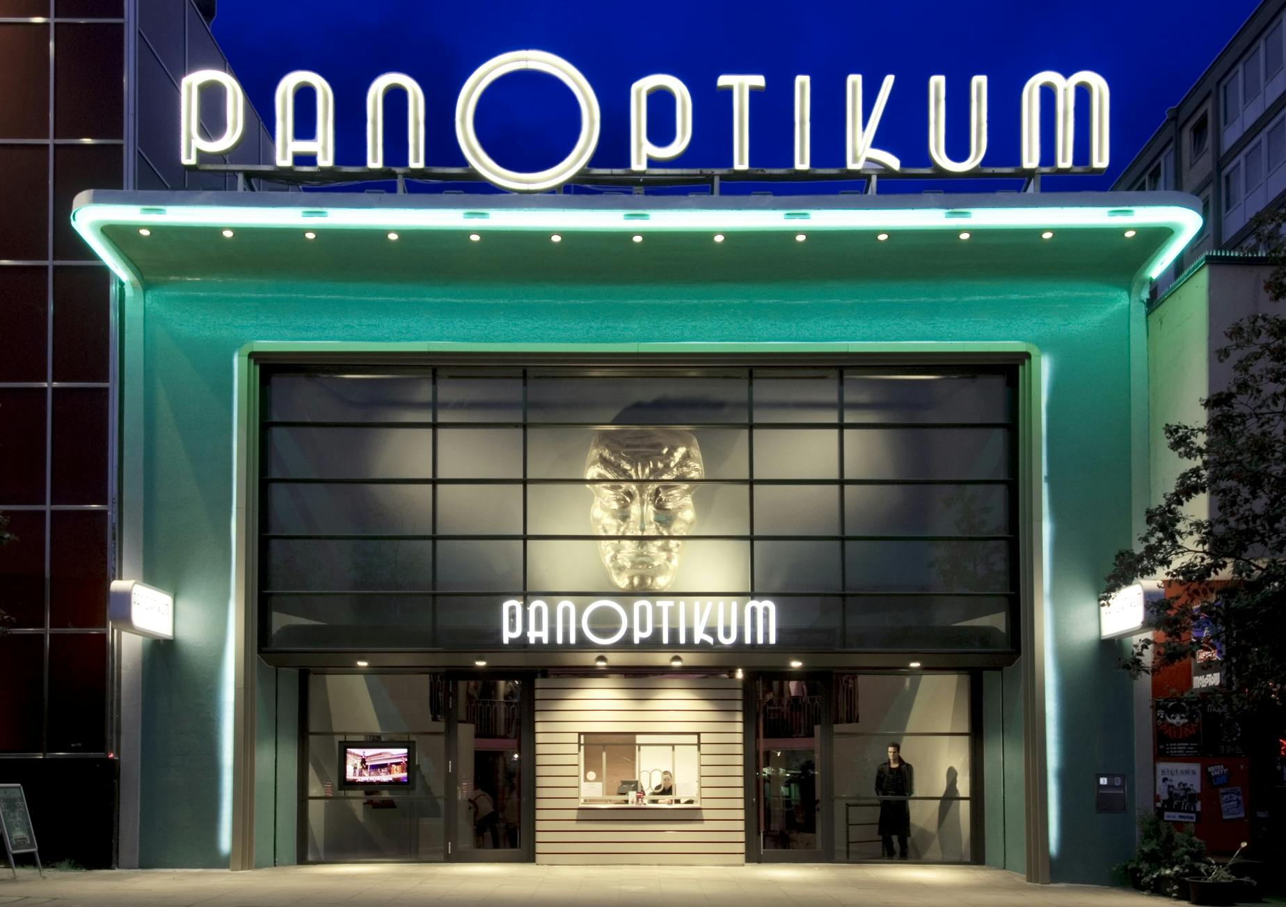 A building entrance with neon "PANOPTIKUM" sign at the top and a large metallic face above the doors, illuminated at night.