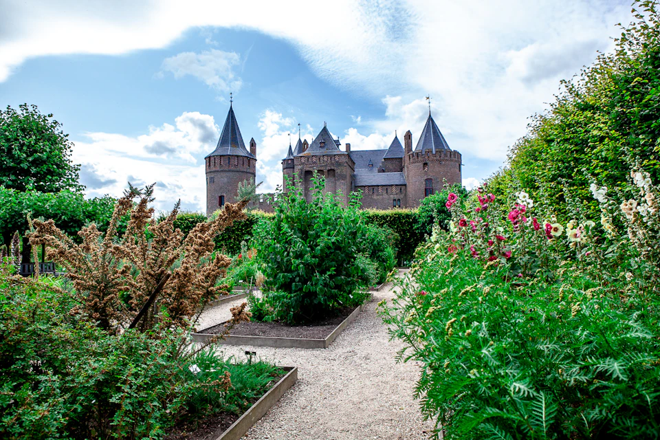 A large brick castle with several towers, seen from a garden filled with various plants and flowers under a partly cloudy sky.