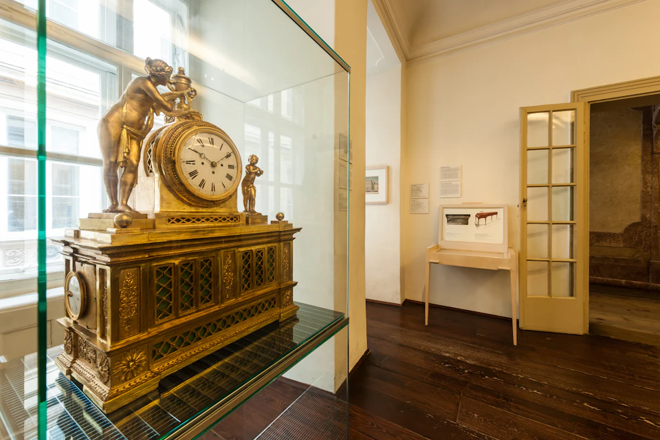 A gold ornamental clock with figurines on display in a well-lit room with wooden floors and framed pictures on the walls.
