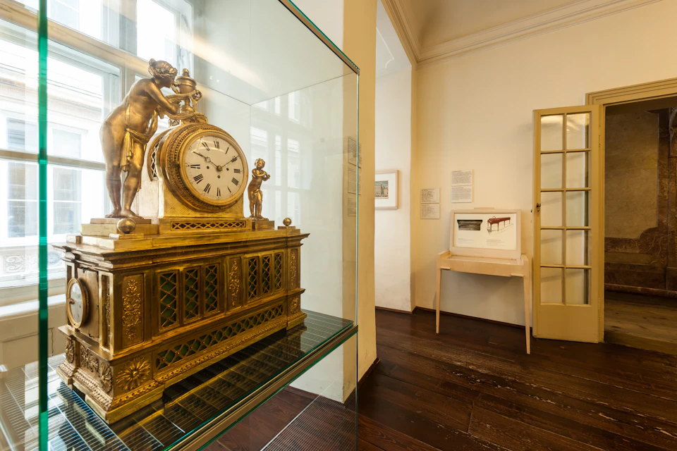 A gold ornamental clock with figurines on display in a well-lit room with wooden floors and framed pictures on the walls.