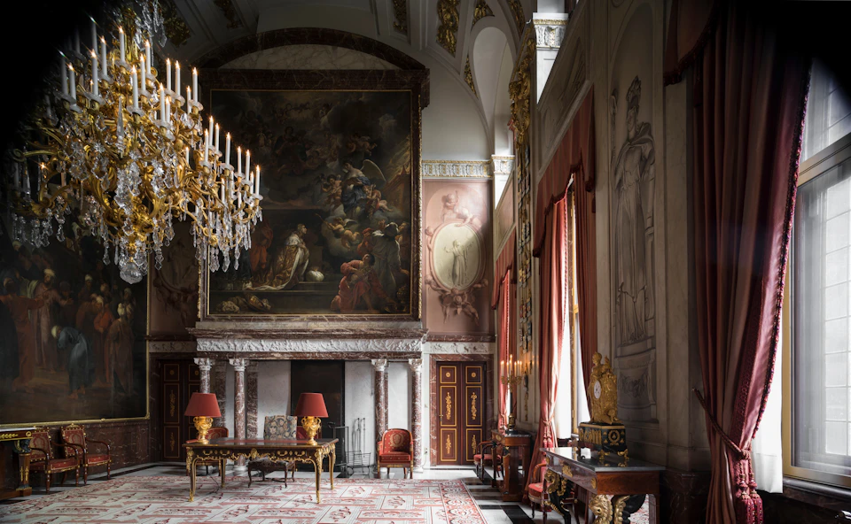 Ornate room with a large painting above a marble fireplace, chandeliers, red curtains, luxurious furniture, and detailed wall carvings.