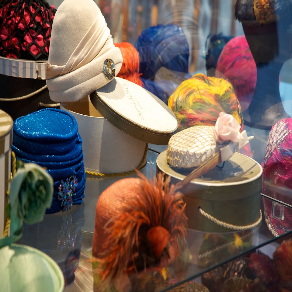 Decorative hats in various colors and styles displayed in a shop window, some resting on round hat boxes.