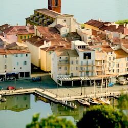 En kuststad med ett klocktorn, omgivande byggnader, kaféer vid vattnet, båtar vid bryggan och människor som går längs kajen.