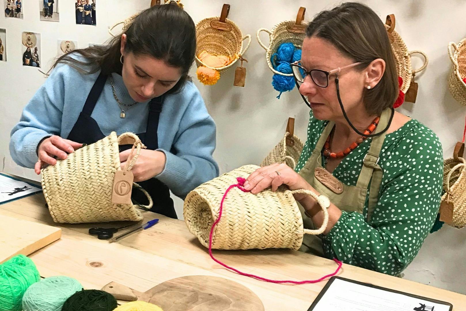 A woman wearing glasses and a green polka-dot shirt weaves red yarn into a straw basket, surrounded by hanging baskets.