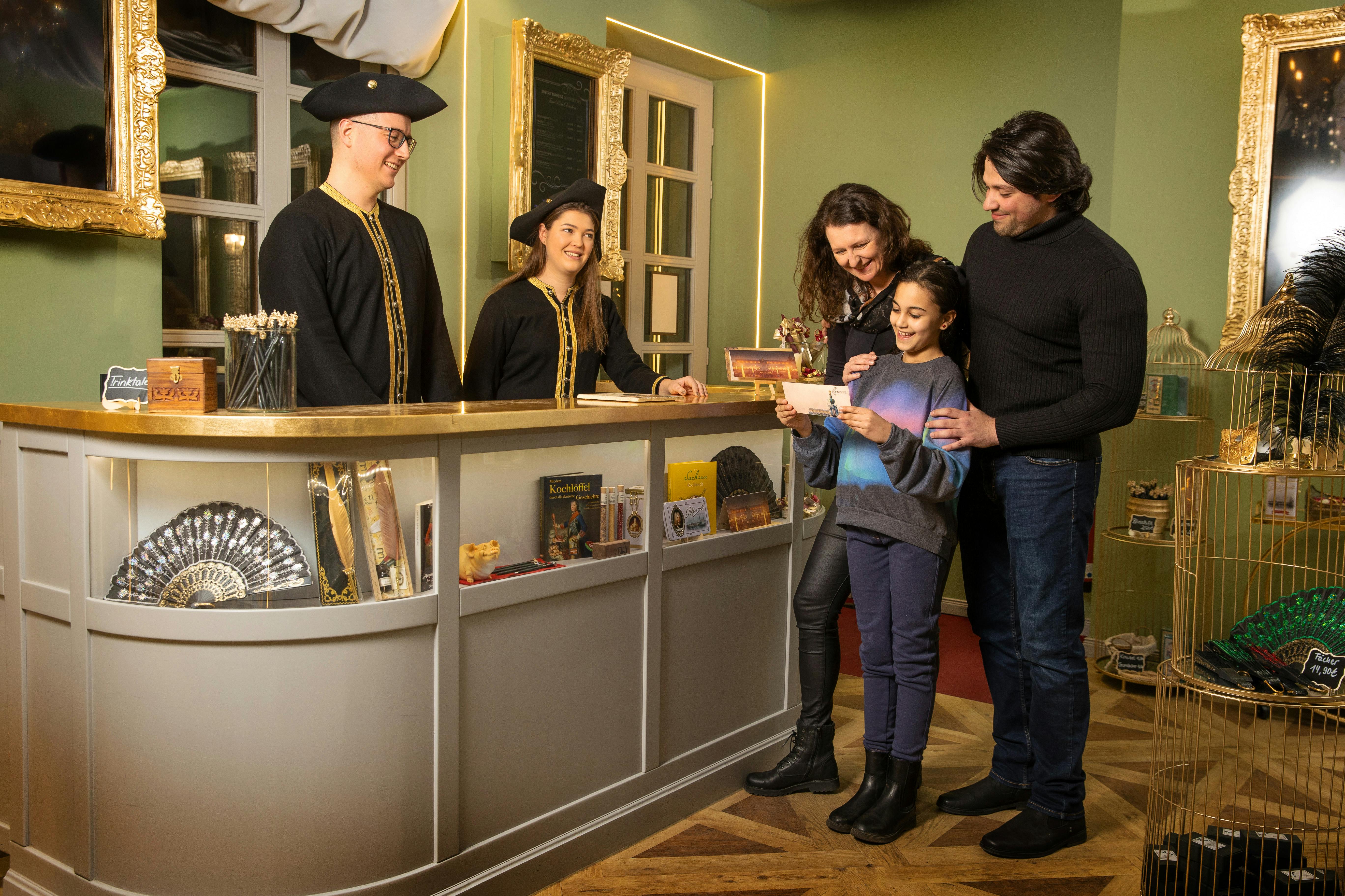 Tři zaměstnanci v černých a zlatých uniformách za pultem slouží usměvavé rodině, která si prohlíží předměty a drží knihu v útulném pokoji.