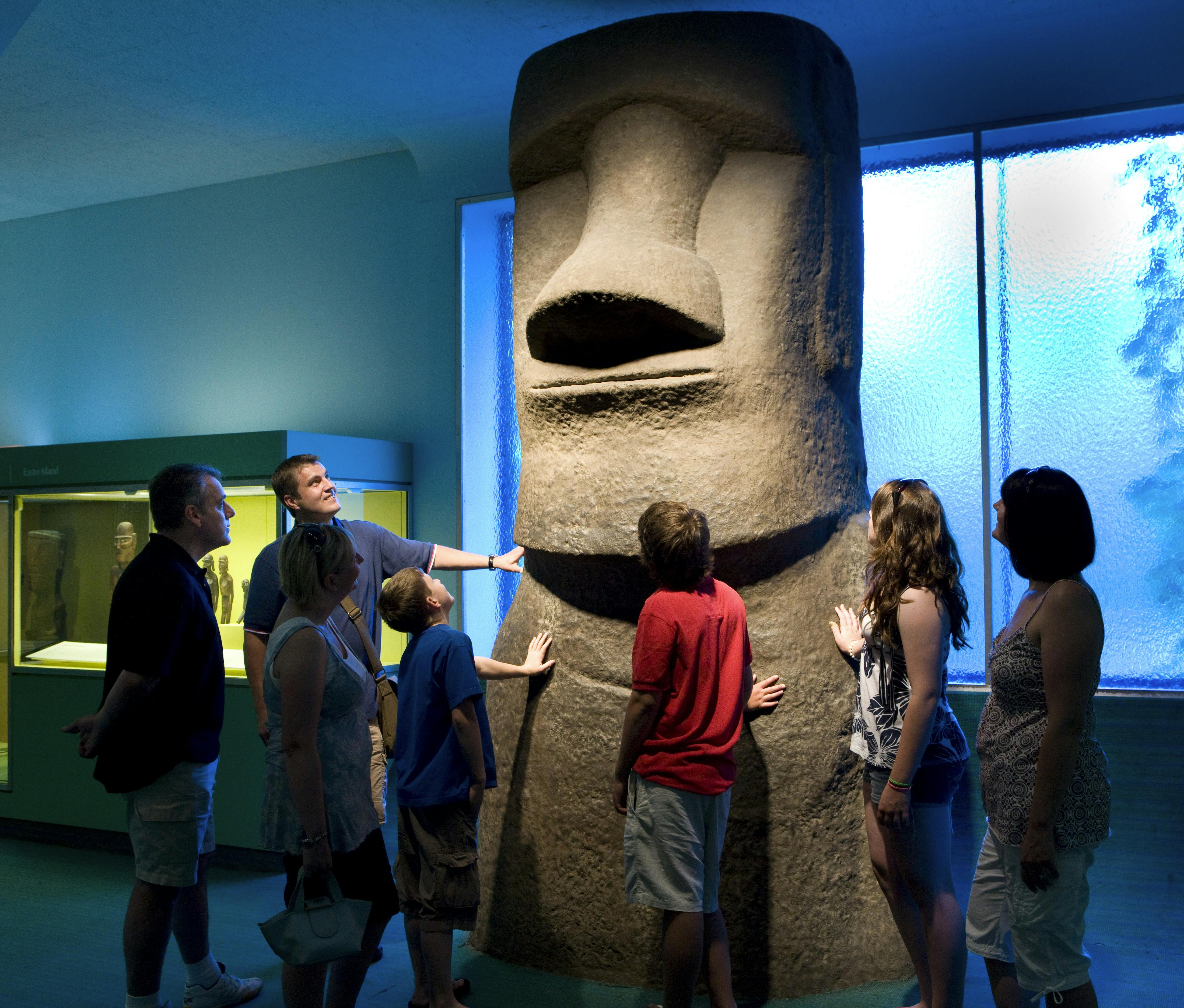 A group of people in a museum admire a tall stone statue resembling a large human head with prominent features.