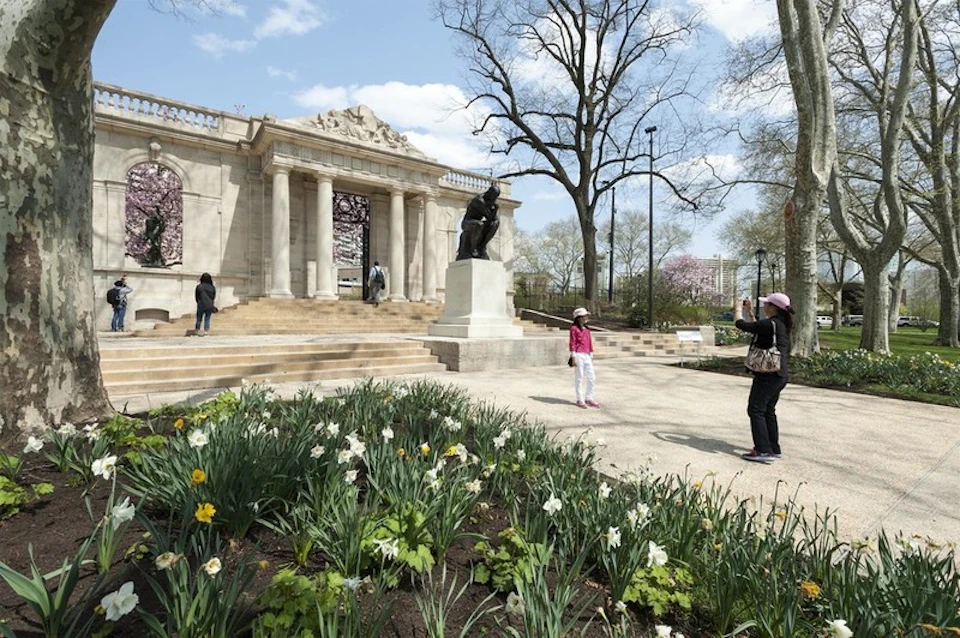 Two people pose for a photo near a statue in front of a large historic building with columns, surrounded by blooming flowers and trees.