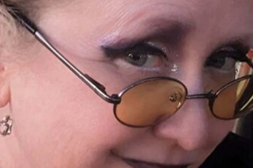 A person with styled blonde hair, wearing yellow-tinted glasses, black attire, and dark makeup, looking at the camera.