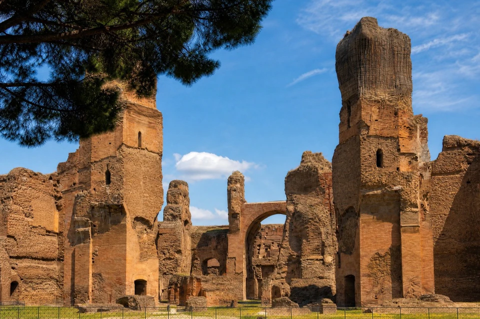 Ancient ruins of a large brick structure with tall walls and arches, under a bright blue sky with scattered clouds.