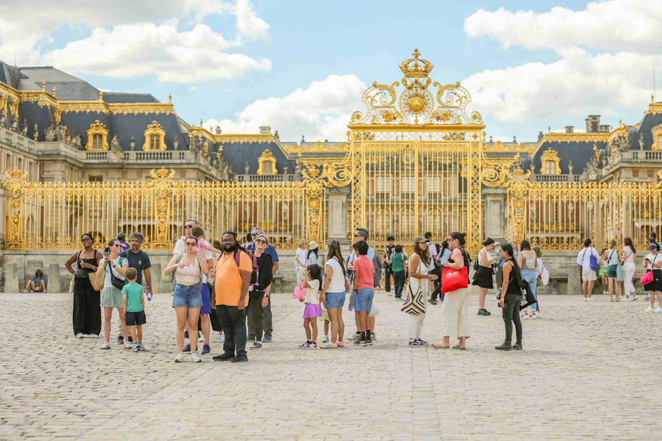 People standing in front of the ornate golden gates of the Palace of Versailles on a sunny day.