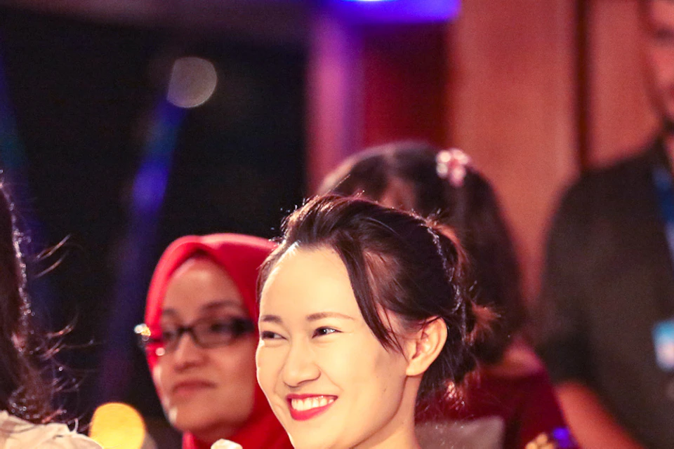 Three women sitting and smiling at an event. One holds a red phone. Other attendees are visible in the background.