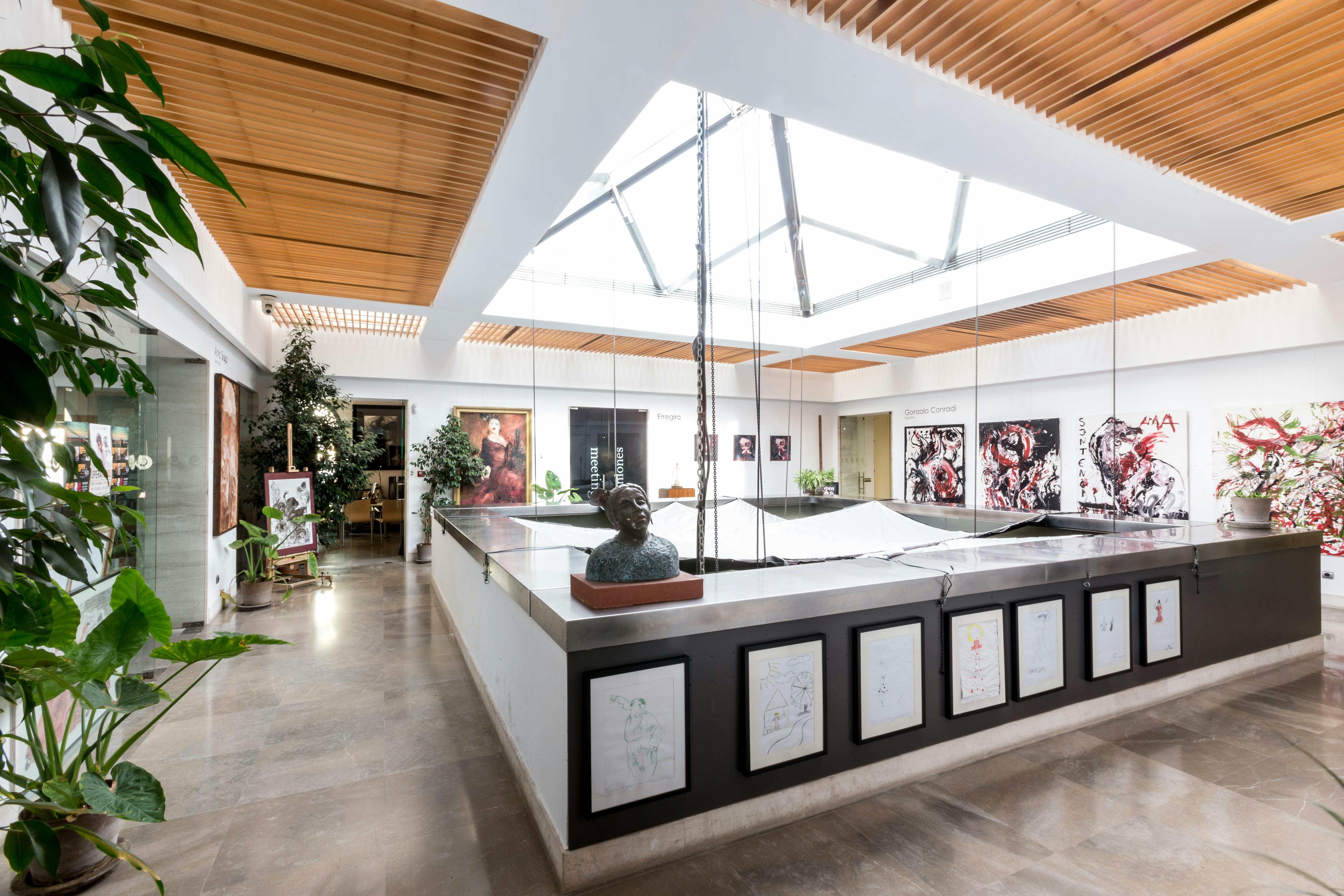 Een moderne kunstgalerie met dakramen, ingelijste kunstwerken, beelden en planten in een ruime, goed verlichte ruimte.