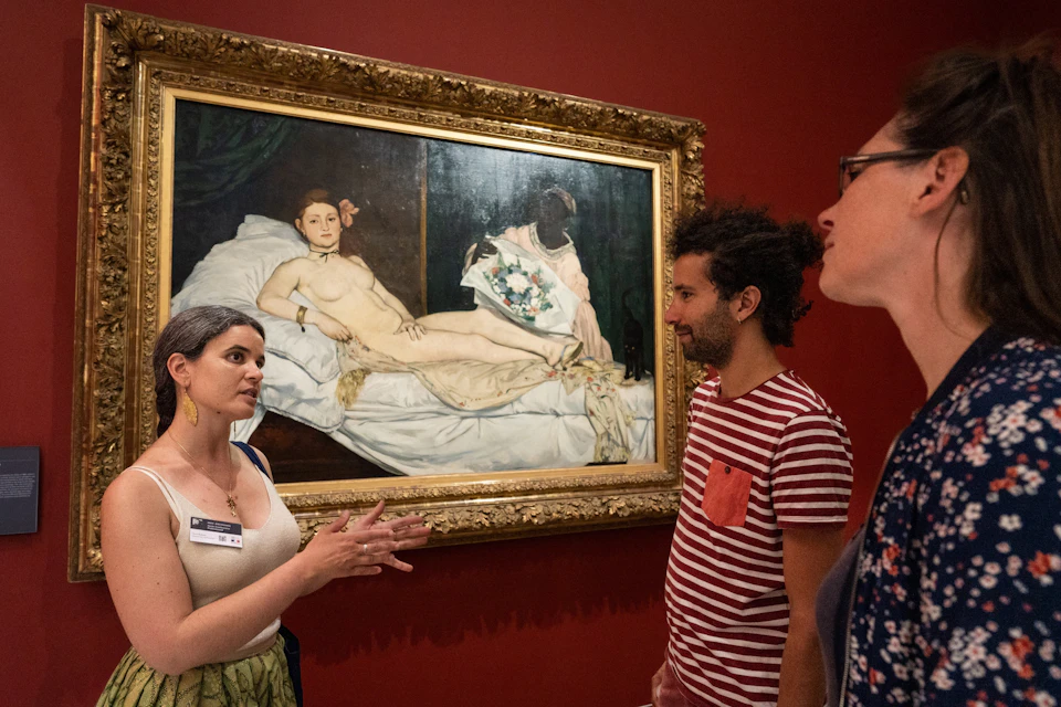 Two people discussing a famous painting of a reclining nude woman in a museum gallery with red walls.