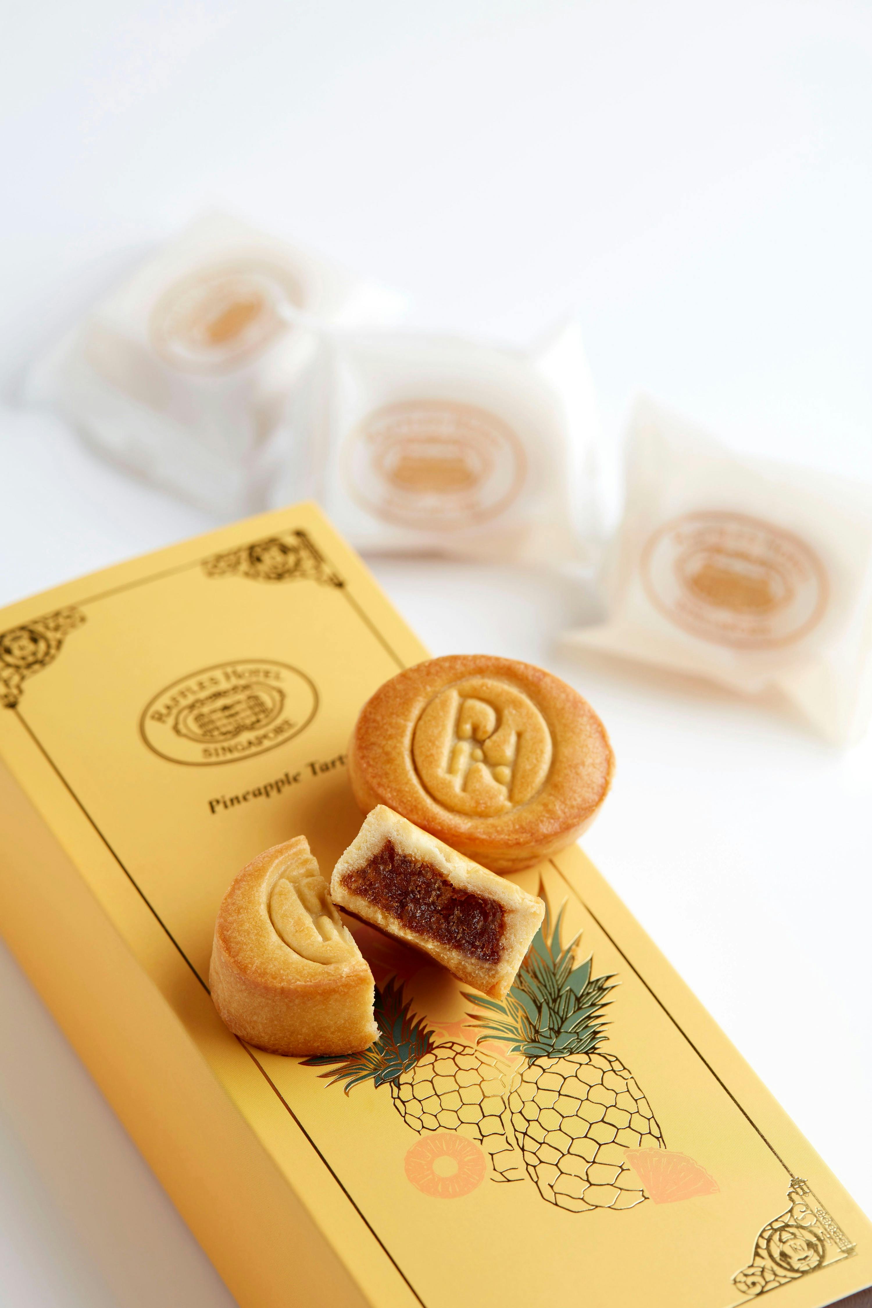 A box of pineapple tarts with two unwrapped tarts, one showing a pineapple filling, and wrapped tarts in the background.