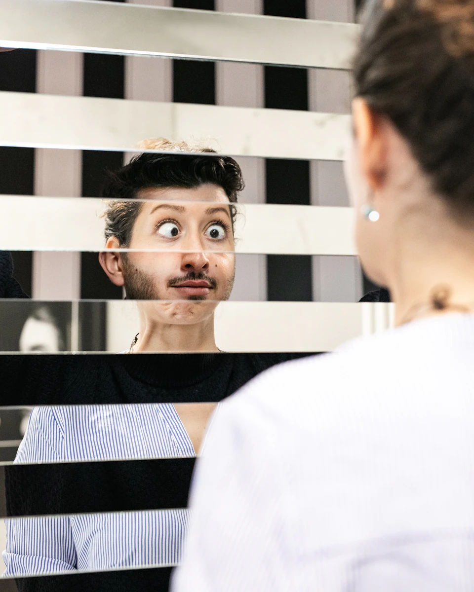 Person with a humorous expression reflecting in a staggered panel mirror, creating a distorted view of their face.