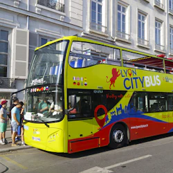En färgglad Lyon City Bus med passagerare som går ombord, parkerad bredvid en byggnad en solig dag.