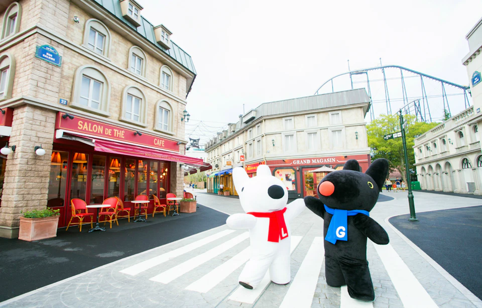 Two mascot characters, one white with a red scarf and one black with a blue scarf, stand on a crosswalk in a quaint street scene.
