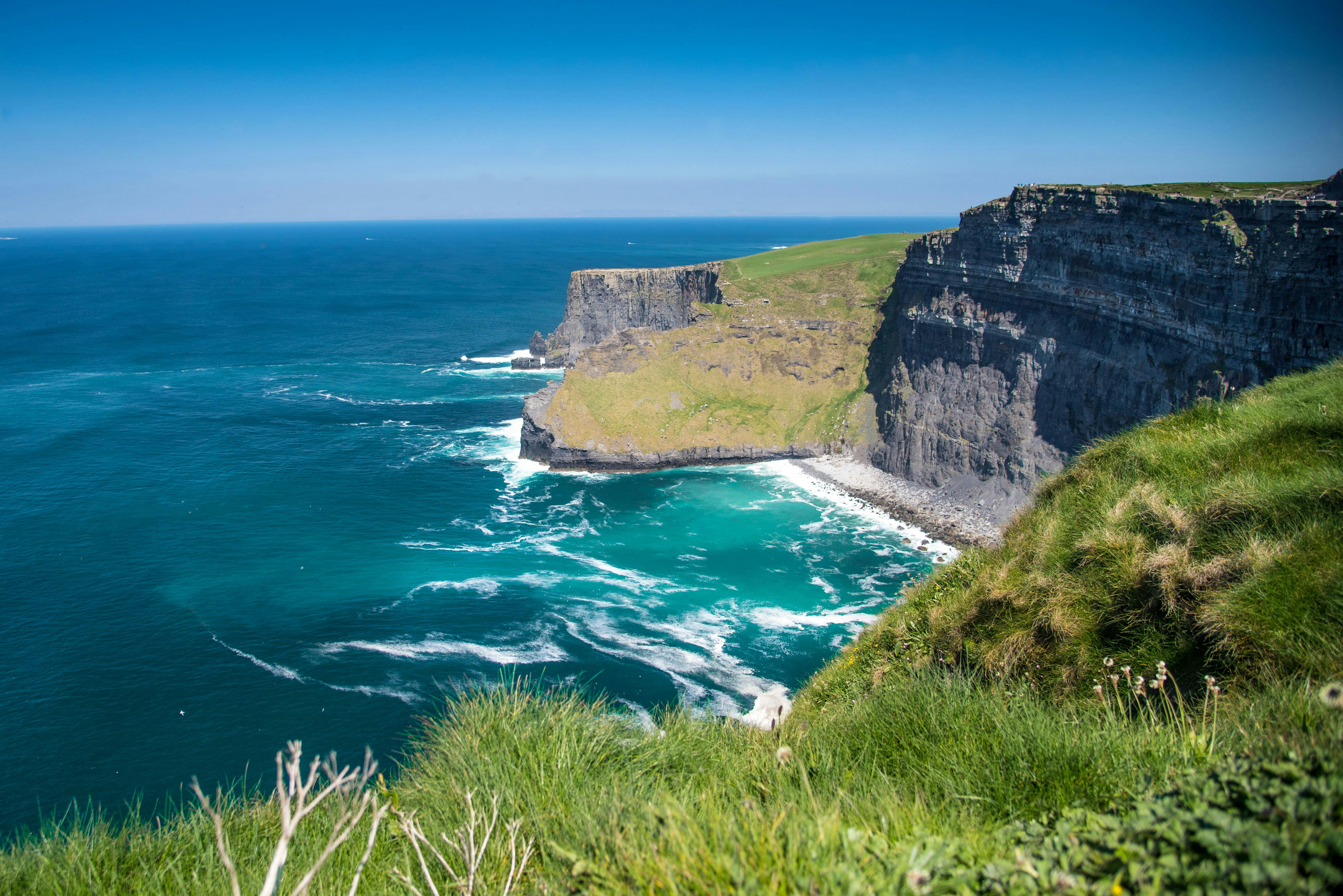 High ocean cliffs with green tops, rugged faces, and crashing waves below under a clear blue sky. Grass in the foreground.