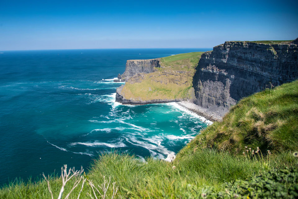 High ocean cliffs with green tops, rugged faces, and crashing waves below under a clear blue sky. Grass in the foreground.