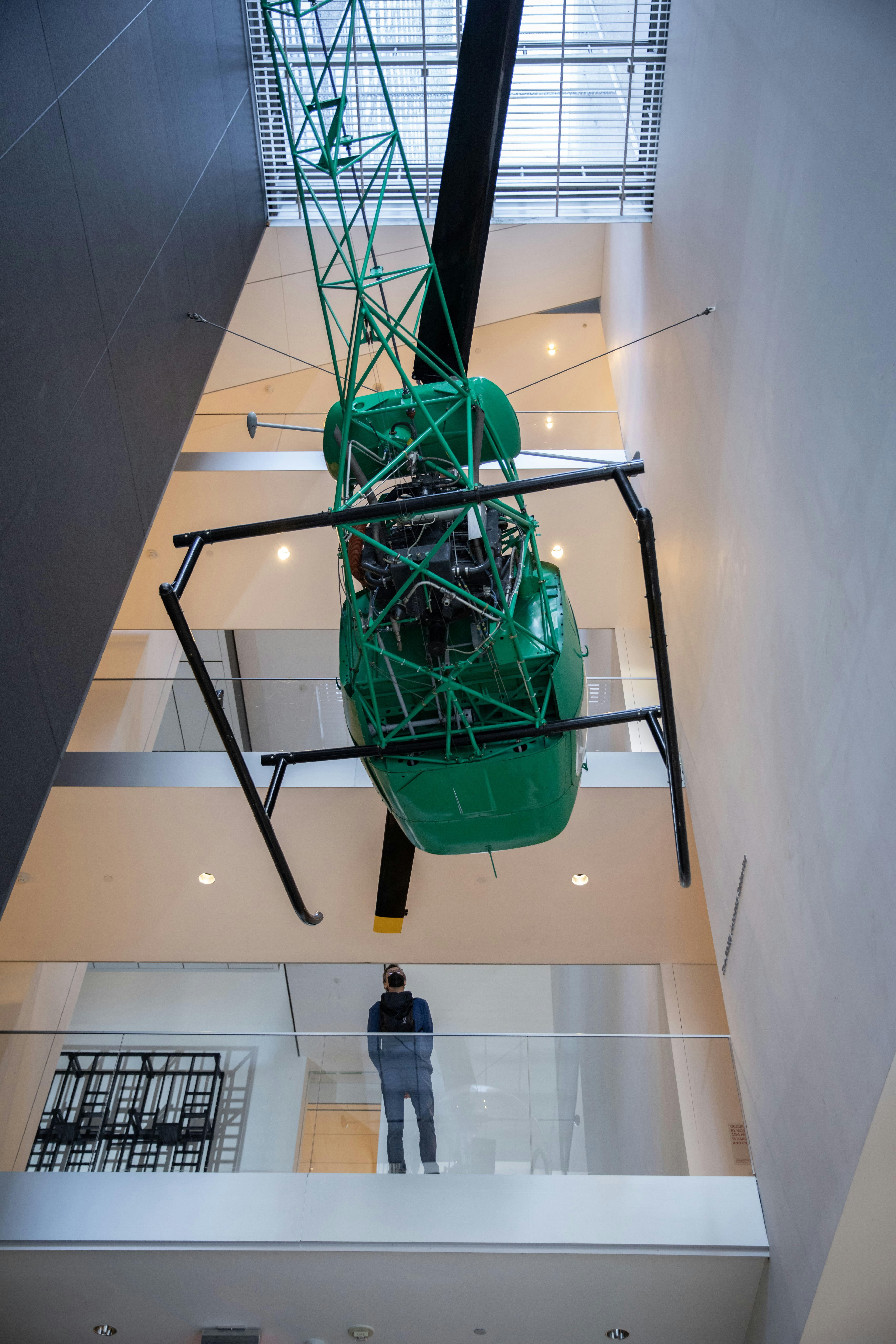 Uma pessoa está num nível inferior, observando um helicóptero verde suspenso do teto no átrio de um edifício moderno.