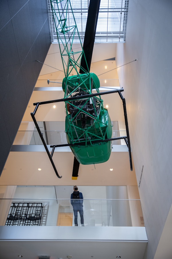 En person står på et lavere niveau og observerer en grøn helikopter ophængt fra loftet i et moderne bygningsatrium.