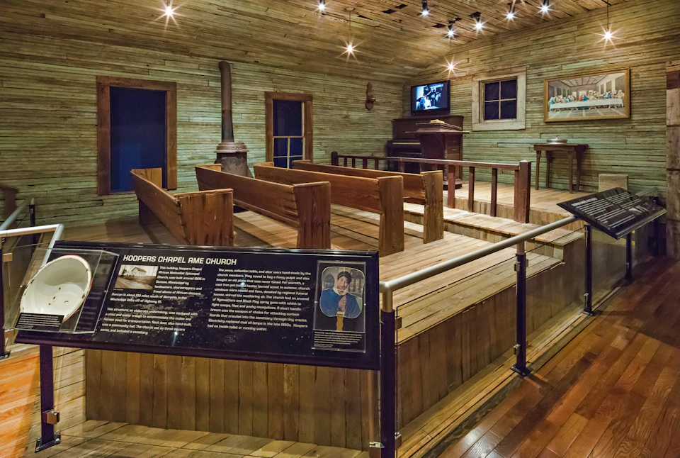 Wooden chapel interior with benches, a wood stove, historical display sign, TV, and piano. Sign labeled "Hoopers Chapel AME Church."