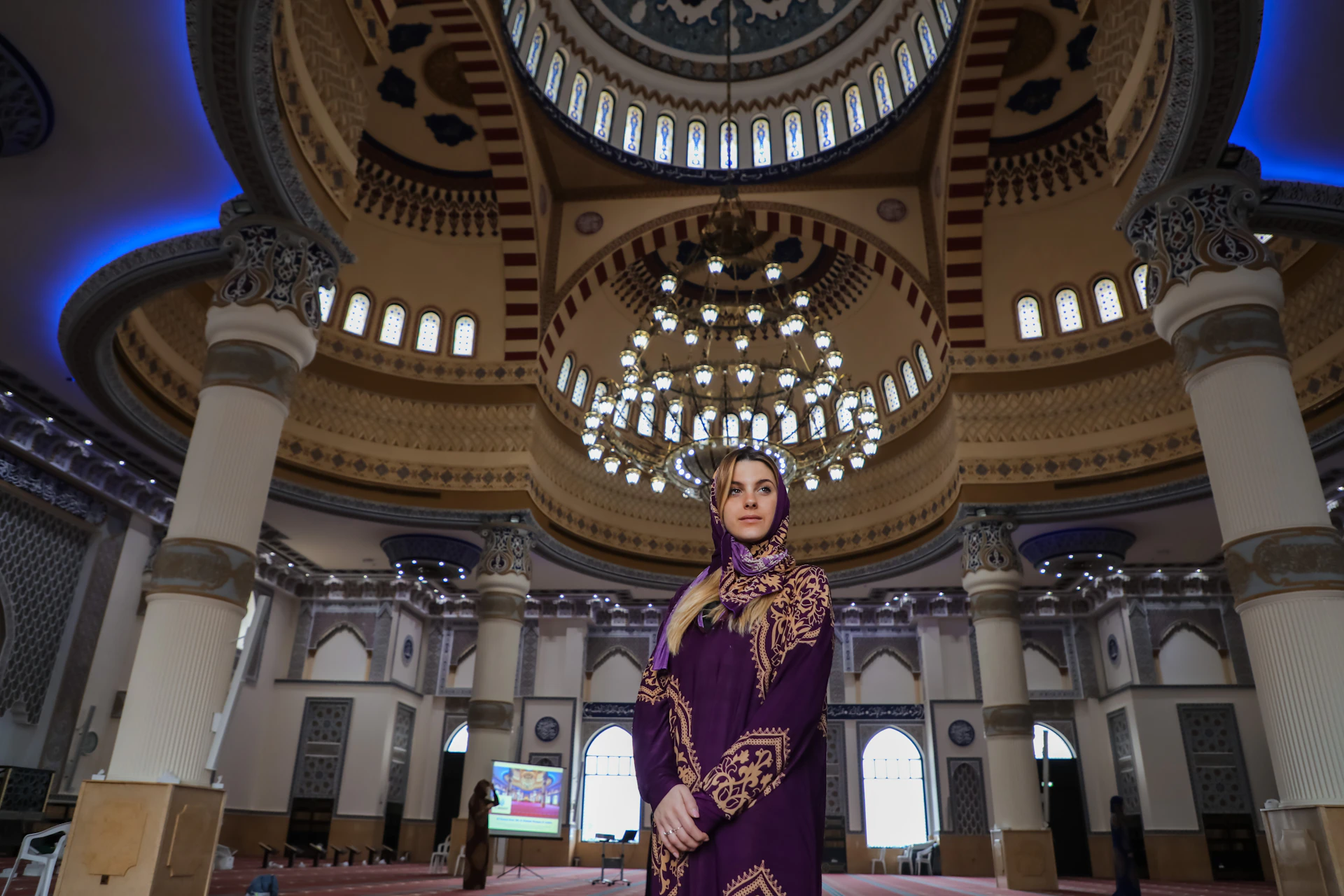 A woman in a purple headscarf stands inside an ornate mosque with a large chandelier and intricate architectural details.