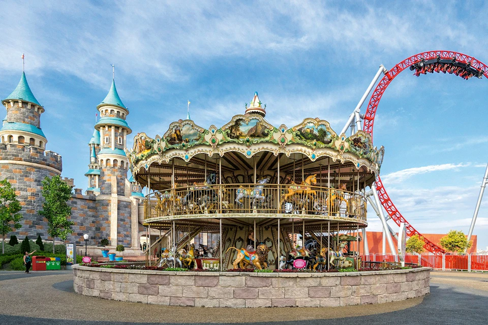 A two-story carousel with painted horses stands in front of a castle-like building and a red roller coaster track under a blue sky.