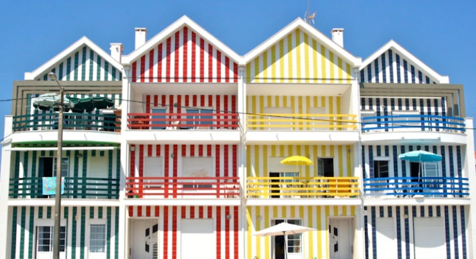 Two adjacent houses with striped facades—one red and white, the other yellow and white—each with balconies and a pitched roof.