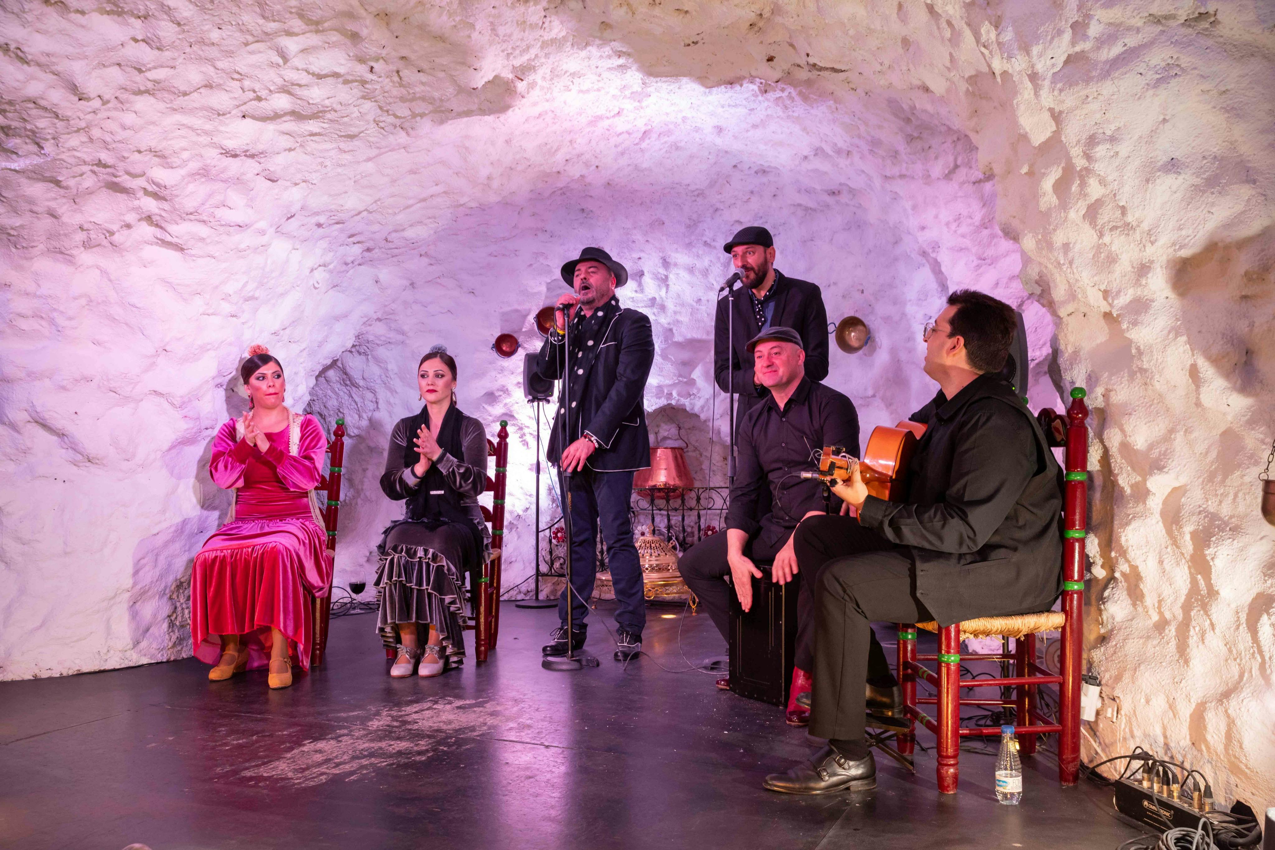 A group of performers, including singers and musicians, in a cave-like setting, with props and lighting enhancing the ambiance.
