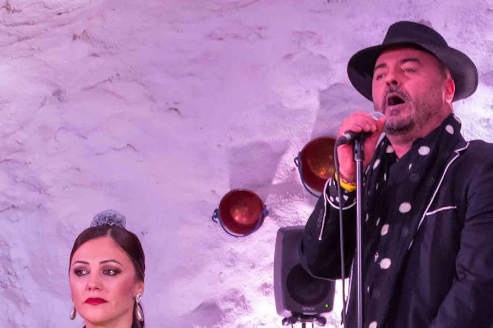A group of performers, including singers and musicians, in a cave-like setting, with props and lighting enhancing the ambiance.