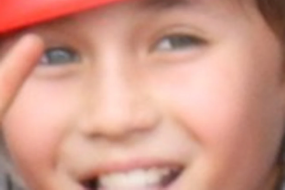 Child climbing a rock wall wearing a red helmet, smiling and making a peace sign with one hand while holding onto grips.