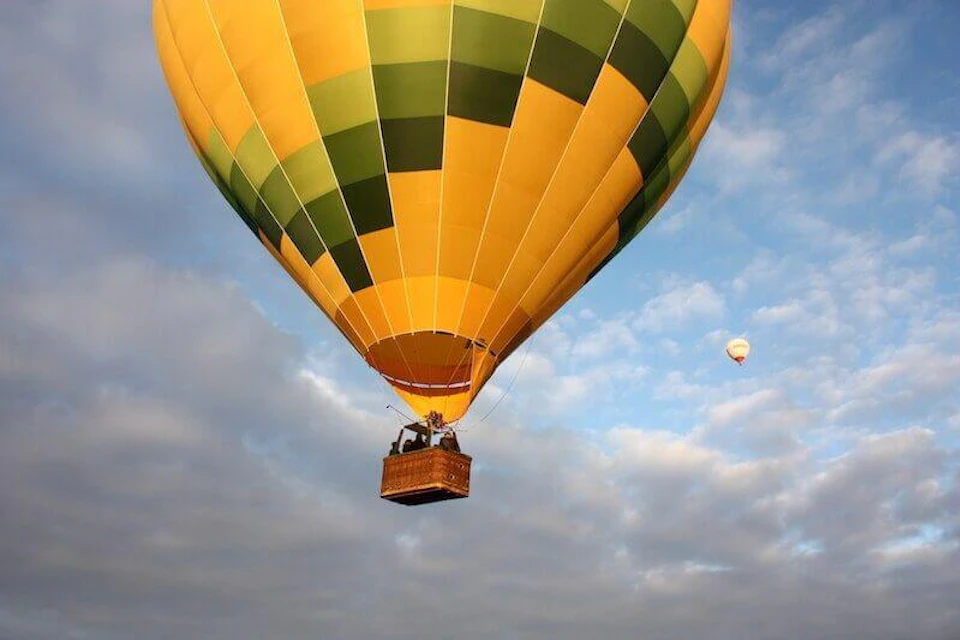 A large yellow and green hot air balloon with a basket floats against a partly cloudy sky; a smaller balloon is in the distance.