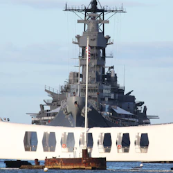 A large battleship is docked behind a white memorial structure with a visible American flag in the center.